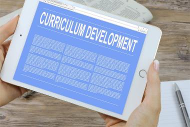 A person holding a white tablet displaying a blue screen with the words “Curriculum Development” in large white text at the top, followed by several blocks of smaller, unreadable text. The tablet rests on a wooden surface with a notebook and a pen nearby.